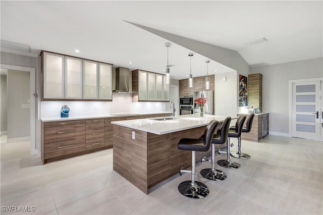 kitchen with a breakfast bar, a sink, light countertops, wall chimney exhaust hood, and a large island with sink