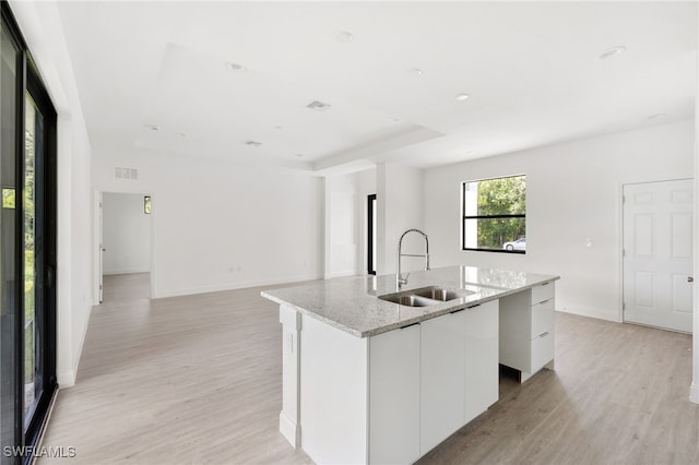 kitchen with white cabinets, an island with sink, light stone countertops, sink, and light wood-type flooring