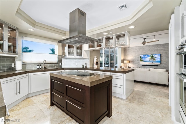 kitchen with a raised ceiling, sink, a kitchen island, stovetop, and island exhaust hood