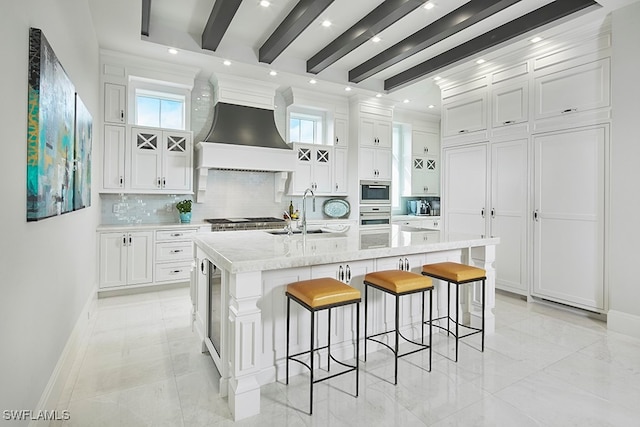 kitchen featuring premium range hood, backsplash, beam ceiling, white cabinets, and a kitchen island with sink