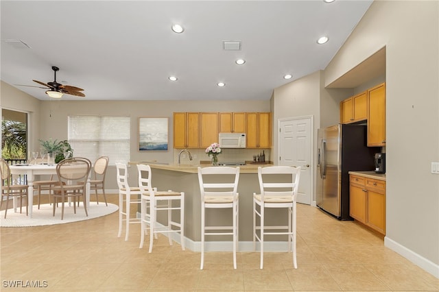 kitchen with ceiling fan, an island with sink, light tile patterned floors, stainless steel refrigerator, and a kitchen bar