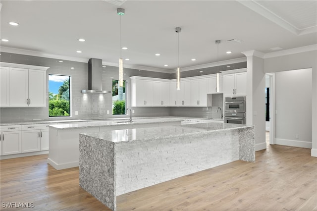 kitchen featuring wall chimney range hood, light stone counters, light wood-type flooring, and a large island with sink