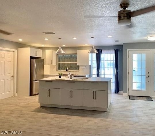 kitchen with stainless steel fridge, a kitchen island with sink, light wood-type flooring, and pendant lighting