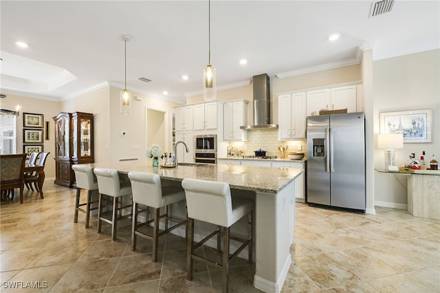 kitchen featuring wall chimney exhaust hood, stainless steel appliances, a kitchen breakfast bar, an island with sink, and white cabinets