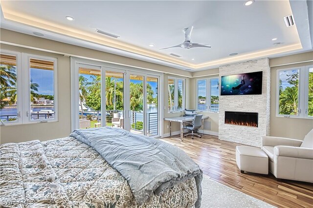 bedroom with ceiling fan, access to outside, a raised ceiling, a stone fireplace, and light hardwood / wood-style flooring