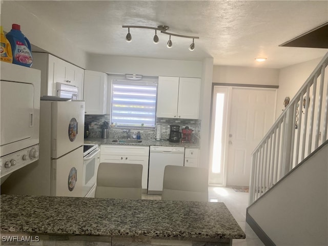 kitchen with dark stone counters, stacked washer and dryer, white cabinets, decorative backsplash, and white appliances