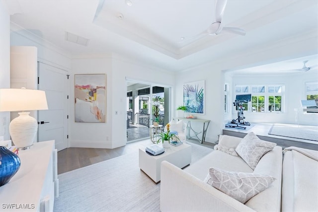 living area featuring light wood finished floors, visible vents, a ceiling fan, ornamental molding, and a tray ceiling