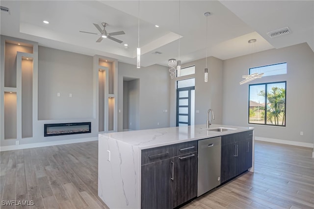 kitchen with dishwasher, a kitchen island with sink, a raised ceiling, sink, and light hardwood / wood-style flooring