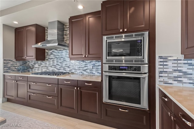 kitchen featuring stainless steel appliances, wall chimney range hood, backsplash, dark brown cabinets, and light tile patterned floors