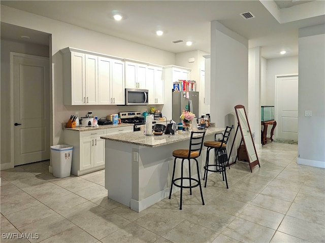 kitchen featuring light tile patterned floors, appliances with stainless steel finishes, white cabinetry, light stone countertops, and a kitchen bar