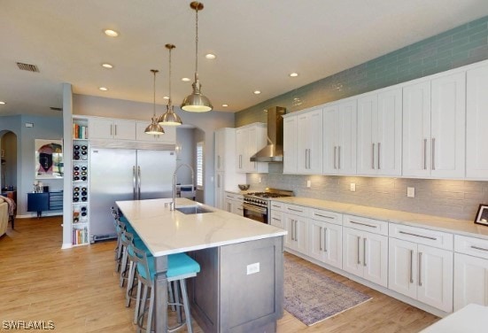 kitchen with wall chimney exhaust hood, a kitchen island with sink, sink, appliances with stainless steel finishes, and white cabinetry