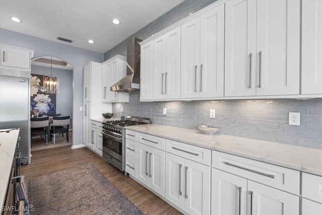 kitchen featuring arched walkways, premium appliances, wood finished floors, white cabinetry, and wall chimney exhaust hood