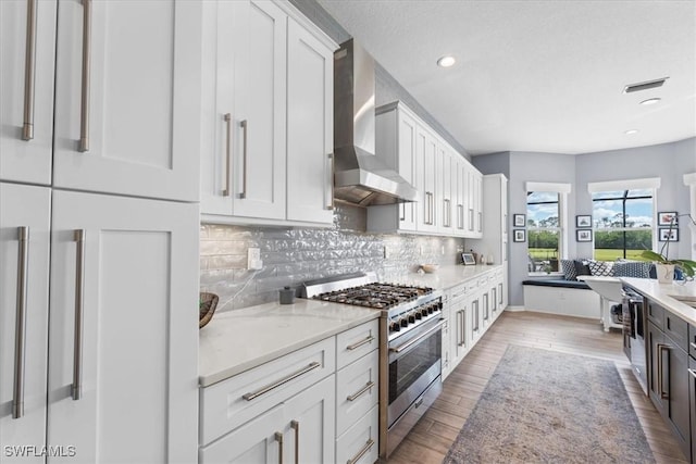 kitchen featuring stainless steel stove, wood finished floors, white cabinetry, wall chimney range hood, and decorative backsplash