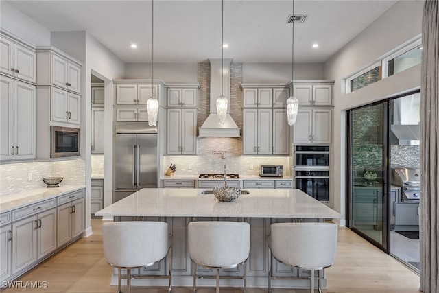 kitchen with light hardwood / wood-style floors, an island with sink, built in appliances, decorative light fixtures, and wall chimney range hood