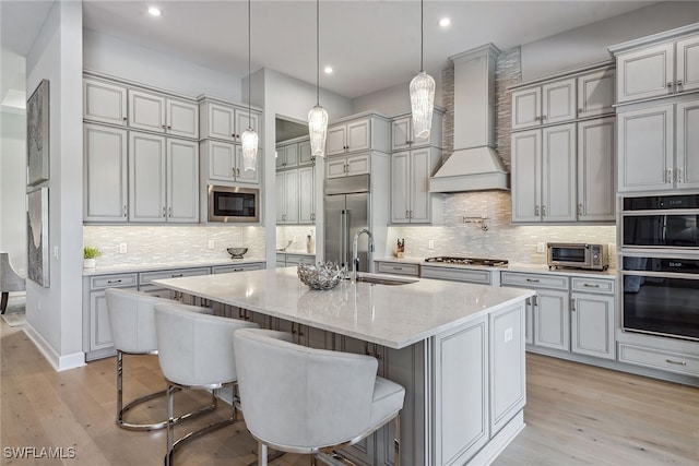 kitchen with custom exhaust hood, an island with sink, sink, built in appliances, and light wood-type flooring