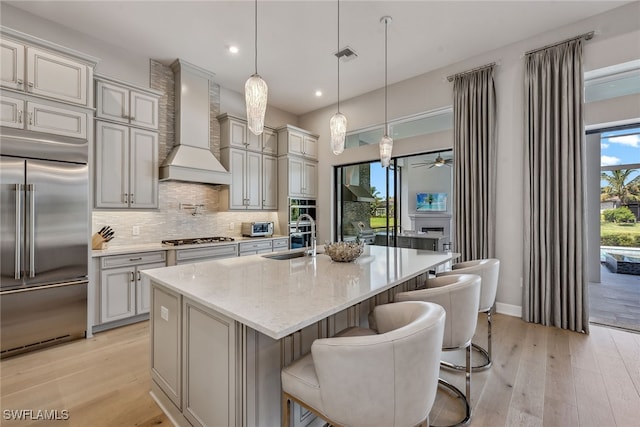 kitchen with light wood-type flooring, a kitchen island with sink, hanging light fixtures, custom range hood, and appliances with stainless steel finishes