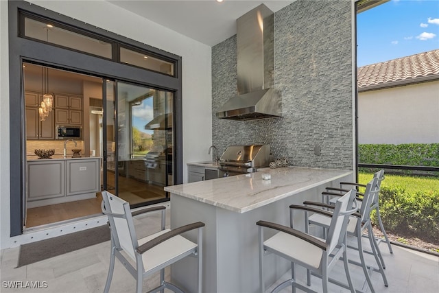 kitchen featuring decorative backsplash, light stone counters, wall chimney exhaust hood, kitchen peninsula, and a kitchen bar