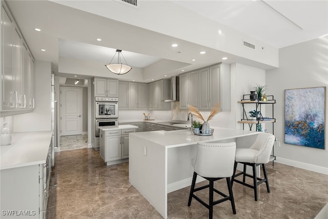 kitchen with hanging light fixtures, wall chimney exhaust hood, double oven, and gray cabinets