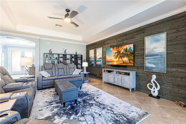 tiled living room with a raised ceiling, wood walls, ceiling fan, and crown molding