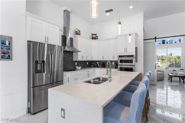 kitchen with an island with sink, stainless steel appliances, sink, wall chimney range hood, and a barn door