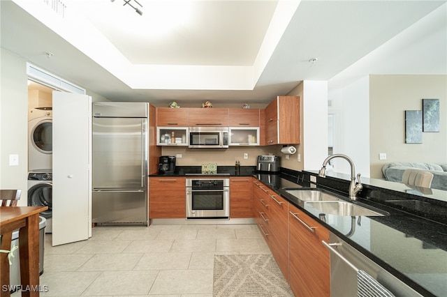 kitchen with sink, dark stone counters, a tray ceiling, appliances with stainless steel finishes, and stacked washer / dryer