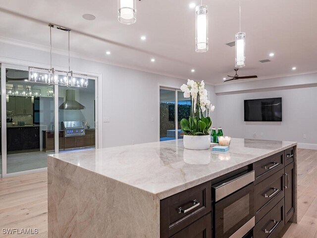 kitchen featuring hanging light fixtures, light stone counters, wall chimney exhaust hood, a center island, and light hardwood / wood-style flooring