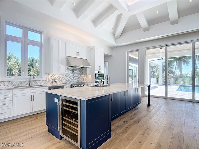 kitchen featuring wine cooler, beamed ceiling, tasteful backsplash, white cabinetry, and a center island with sink