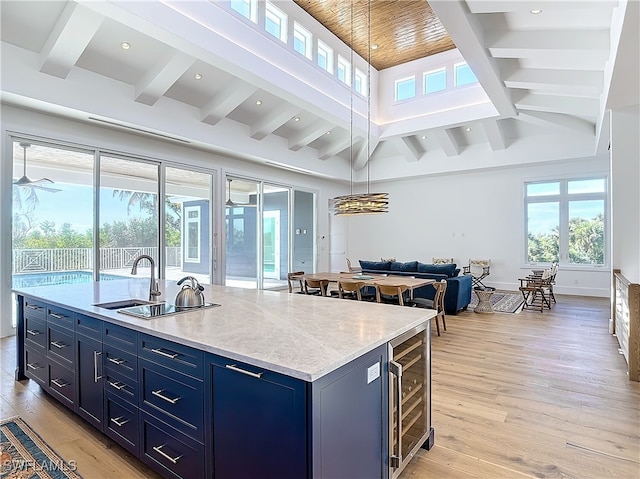 kitchen with beamed ceiling, beverage cooler, a center island with sink, blue cabinetry, and light wood-type flooring