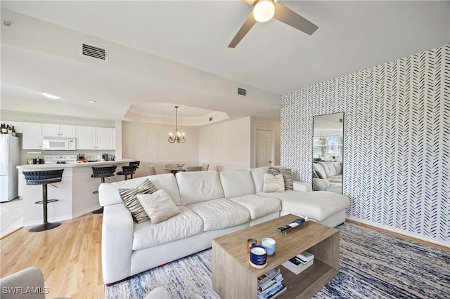 living room featuring ceiling fan with notable chandelier, wallpapered walls, visible vents, and a tray ceiling