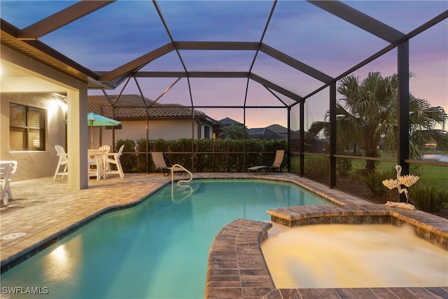 pool at dusk featuring glass enclosure, a patio area, and an in ground hot tub