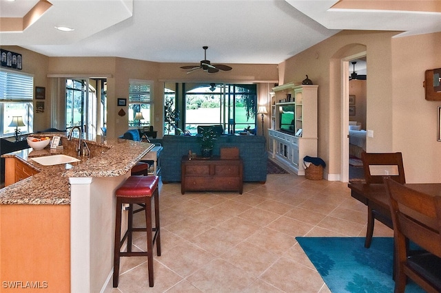 kitchen with light tile patterned flooring, ceiling fan, sink, light stone countertops, and a kitchen bar