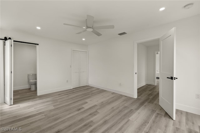 unfurnished bedroom featuring ensuite bathroom, ceiling fan, a barn door, light hardwood / wood-style floors, and a closet