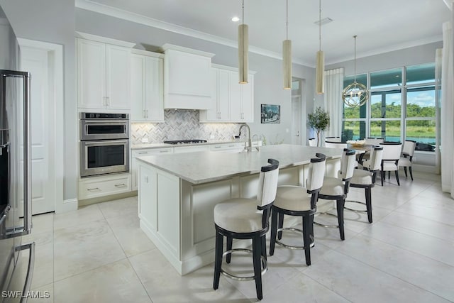 kitchen with stainless steel double oven, an island with sink, hanging light fixtures, light stone countertops, and white cabinets