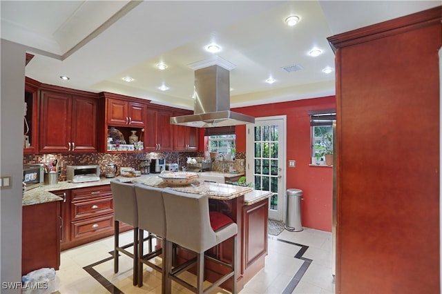 kitchen featuring decorative backsplash, island exhaust hood, light stone counters, and a kitchen bar