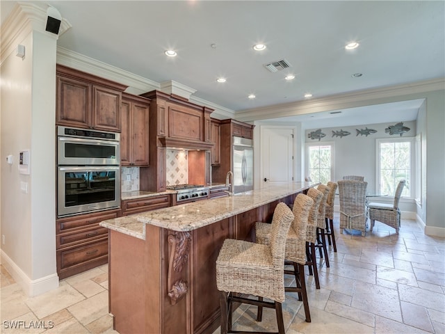 kitchen featuring a large island with sink, backsplash, crown molding, appliances with stainless steel finishes, and light stone counters