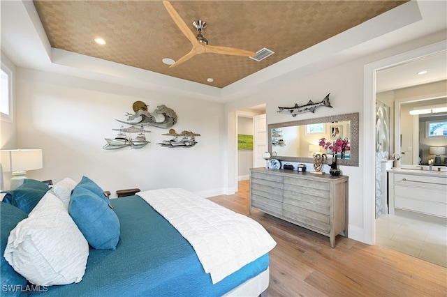 bedroom with ceiling fan, light hardwood / wood-style floors, a raised ceiling, and ensuite bath