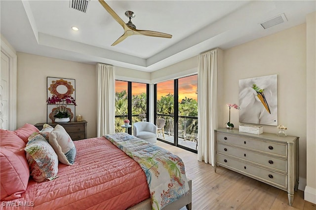 bedroom with light wood-type flooring, access to outside, ceiling fan, and a tray ceiling