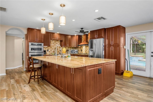 kitchen featuring light hardwood / wood-style floors, a kitchen island with sink, wall chimney range hood, appliances with stainless steel finishes, and decorative light fixtures