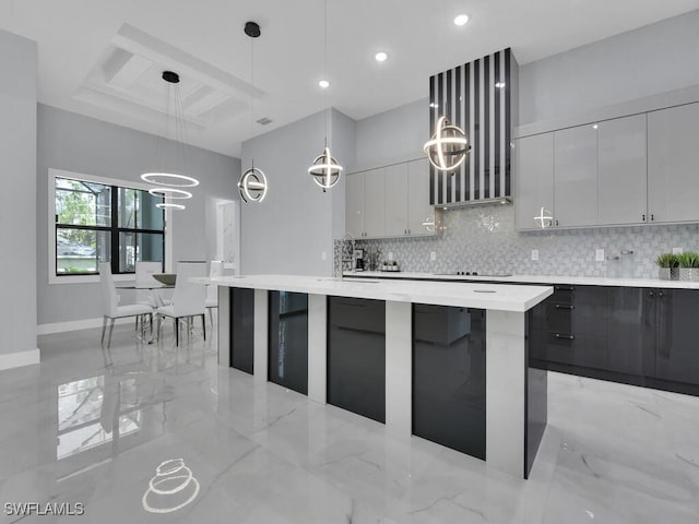 kitchen featuring an island with sink, backsplash, black electric cooktop, decorative light fixtures, and a notable chandelier