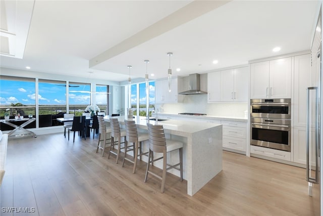 kitchen with stainless steel double oven, wall chimney range hood, an island with sink, hanging light fixtures, and white cabinets