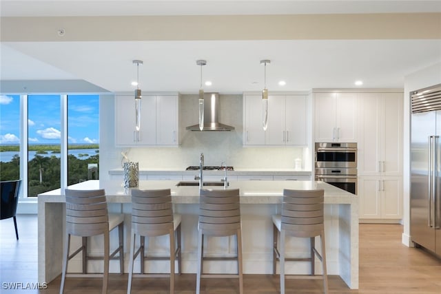 kitchen with white cabinets, wall chimney range hood, an island with sink, and hanging light fixtures