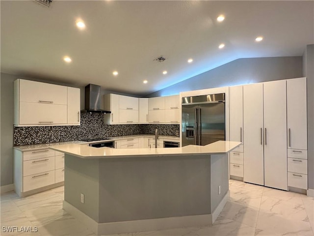 kitchen with lofted ceiling, a large island with sink, wall chimney range hood, and built in fridge