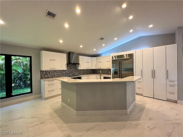 kitchen featuring built in fridge, white cabinets, wall chimney range hood, vaulted ceiling, and a center island with sink