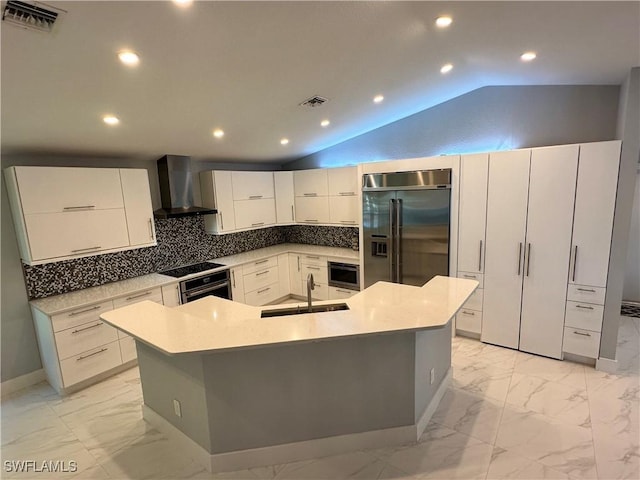 kitchen with wall chimney range hood, a spacious island, built in appliances, vaulted ceiling, and sink