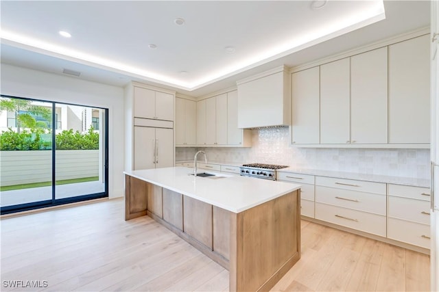 kitchen featuring custom exhaust hood, range, a center island with sink, a tray ceiling, and paneled fridge