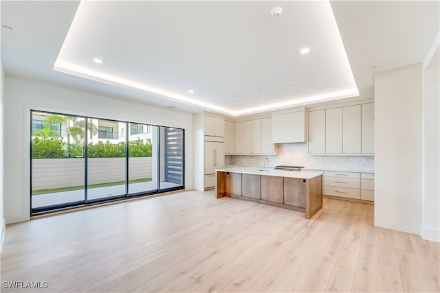 kitchen with a large island with sink, a tray ceiling, tasteful backsplash, and light hardwood / wood-style flooring