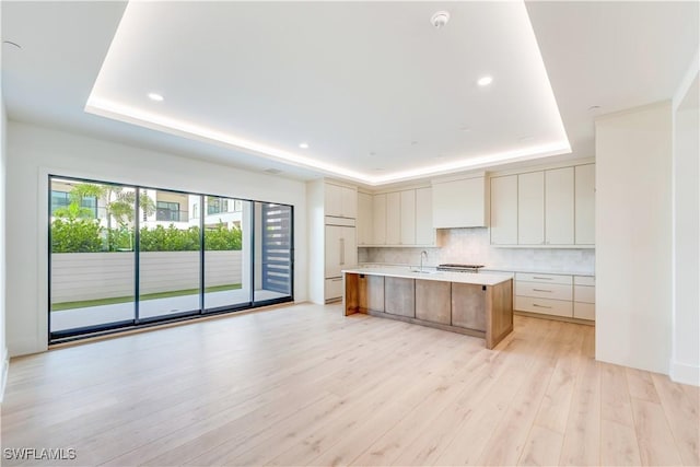 kitchen with backsplash, wall chimney exhaust hood, a tray ceiling, and a large island with sink