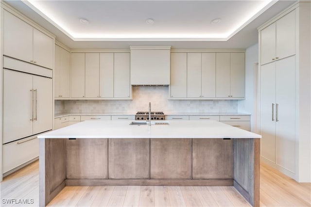 kitchen featuring sink, custom exhaust hood, a raised ceiling, paneled fridge, and a kitchen island with sink