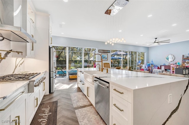 kitchen with ceiling fan, stainless steel appliances, wall chimney range hood, a large island with sink, and light parquet flooring