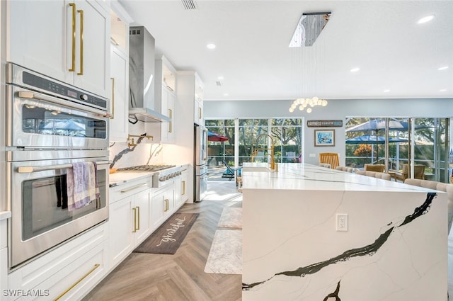 kitchen with light stone countertops, appliances with stainless steel finishes, wall chimney range hood, pendant lighting, and white cabinets